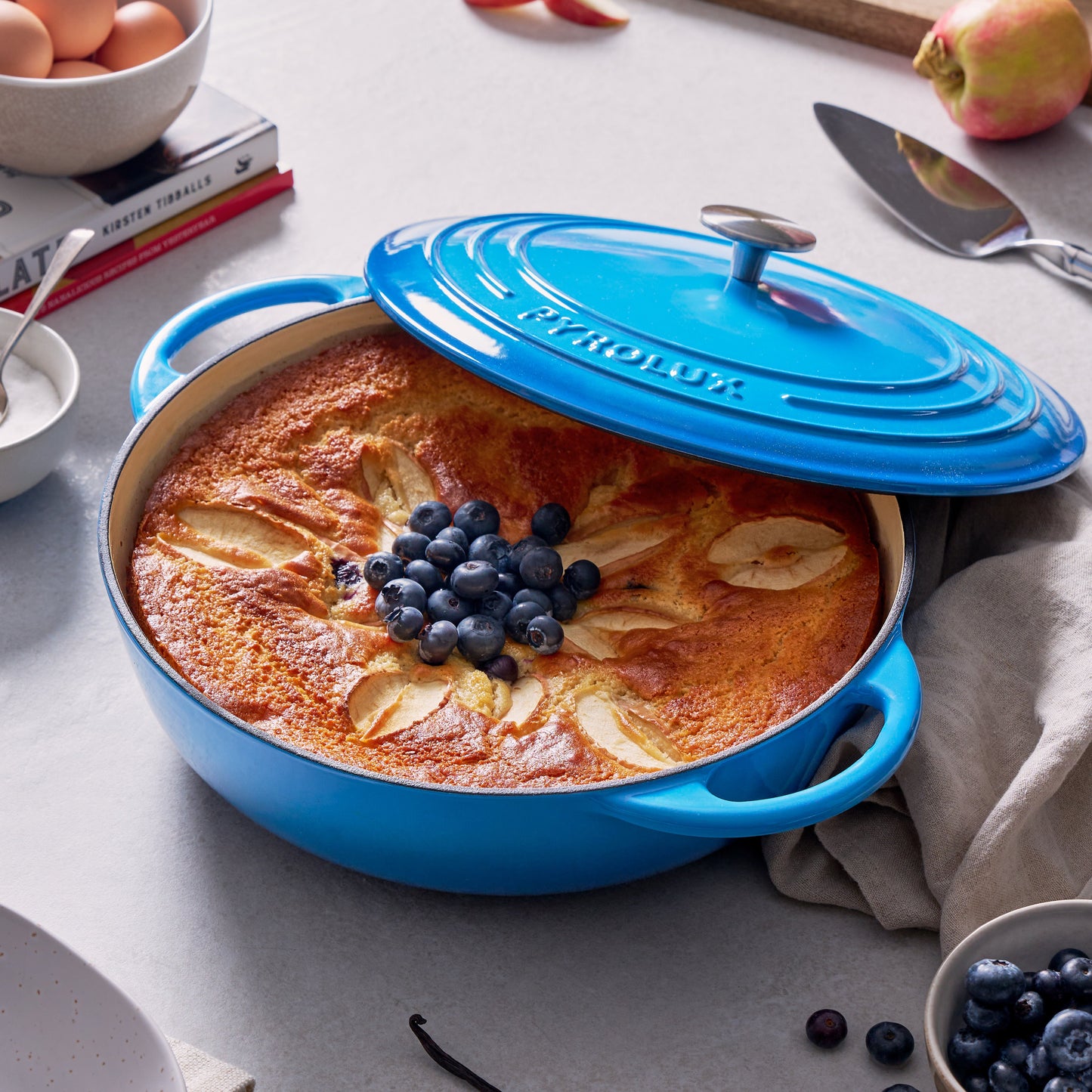 Blue ceramic dish with baked dessert, surrounded by apples, blueberries, and vanilla beans on a light gray surface.
