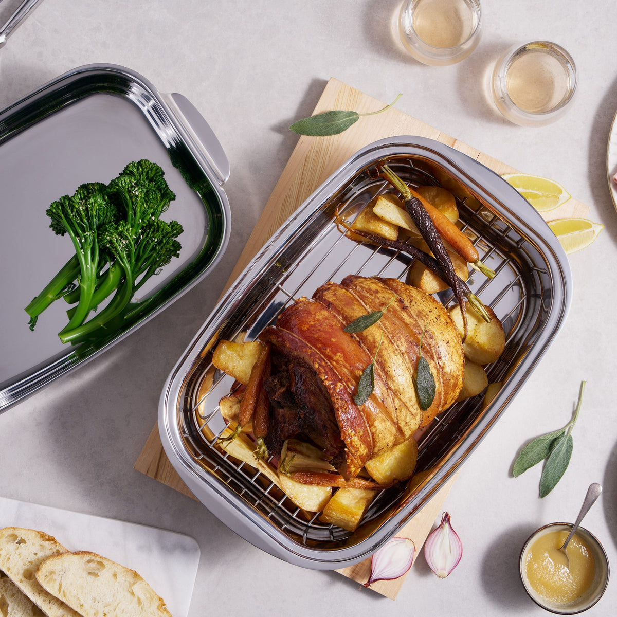 Two rectangular stainless steel roasting pans arranged on a light stone surface, styled with roasted meat, broccoli, bread, plates, and small glasses. No person or animal is present, so this is Product/Packaging rather than Lifestyle, even though it uses a styled tabletop setup.